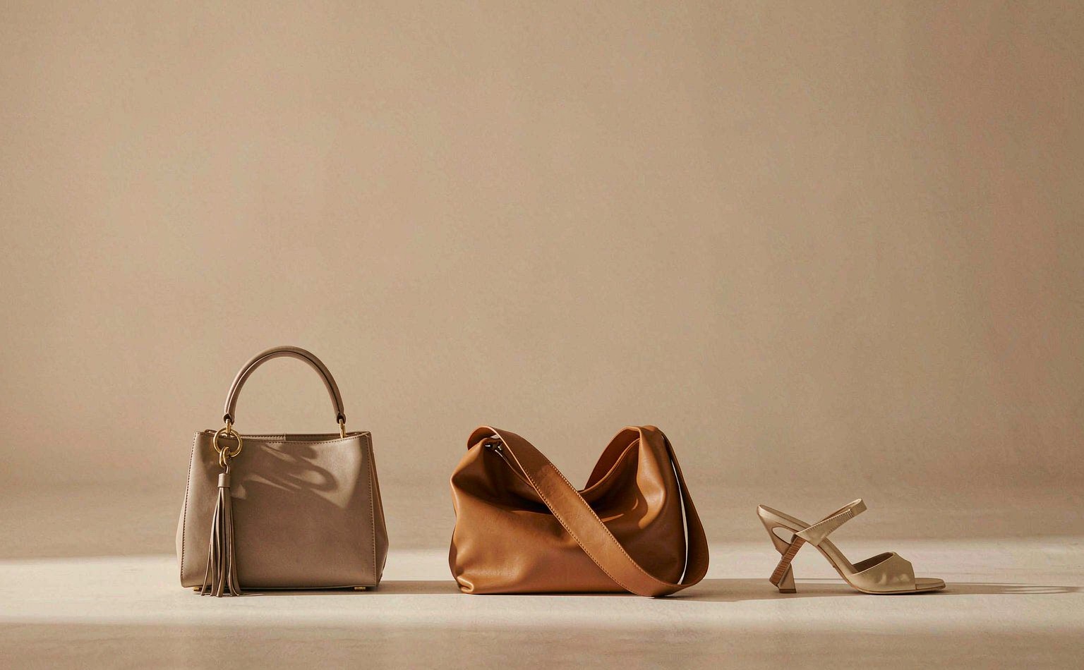 Two Dozoluxe leather handbags and a gold kitten-heel sandal on a warm sand backdrop